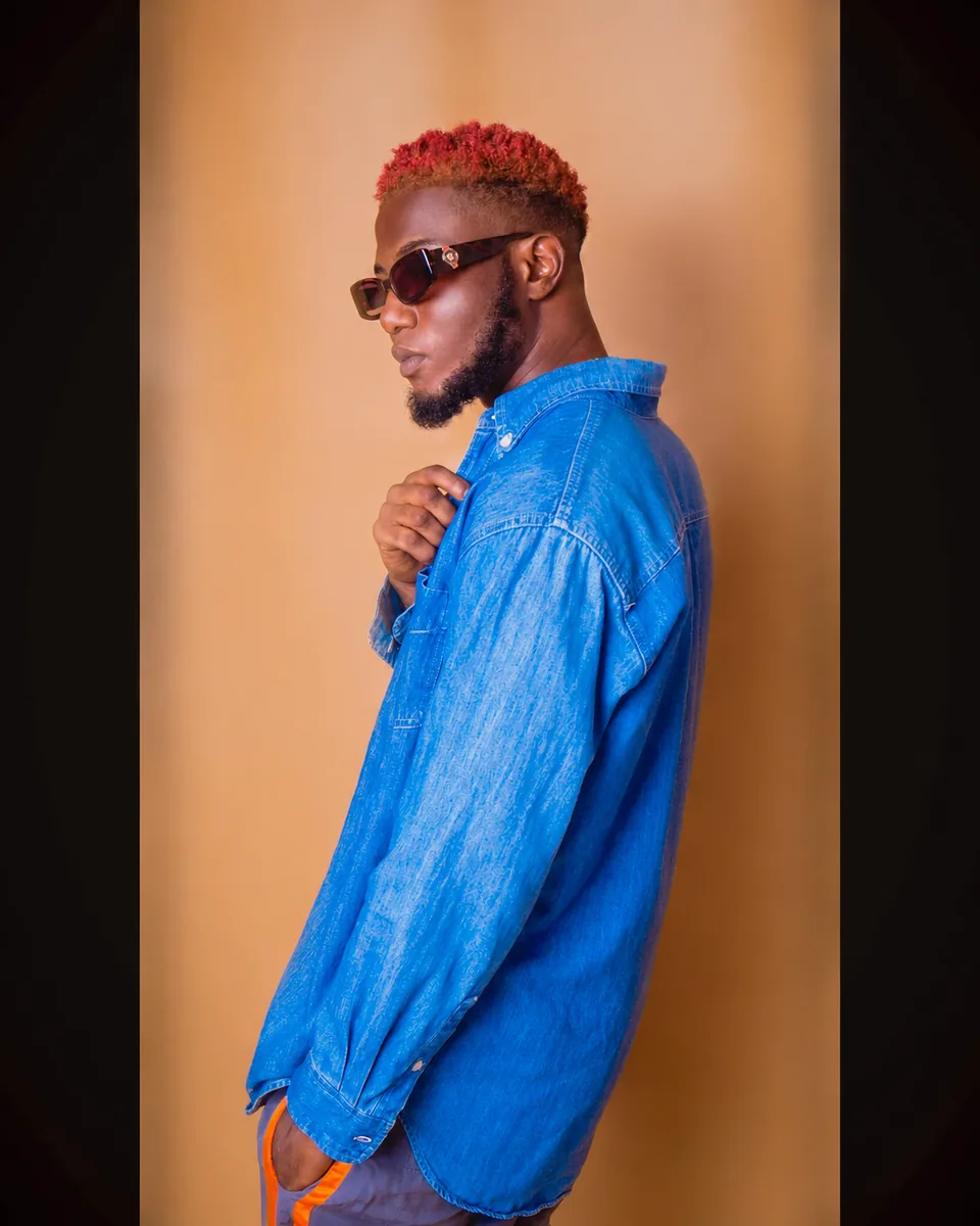 Fashion editorial portrait of a male artist wearing sunglasses and a blue denim shirt against a warm background