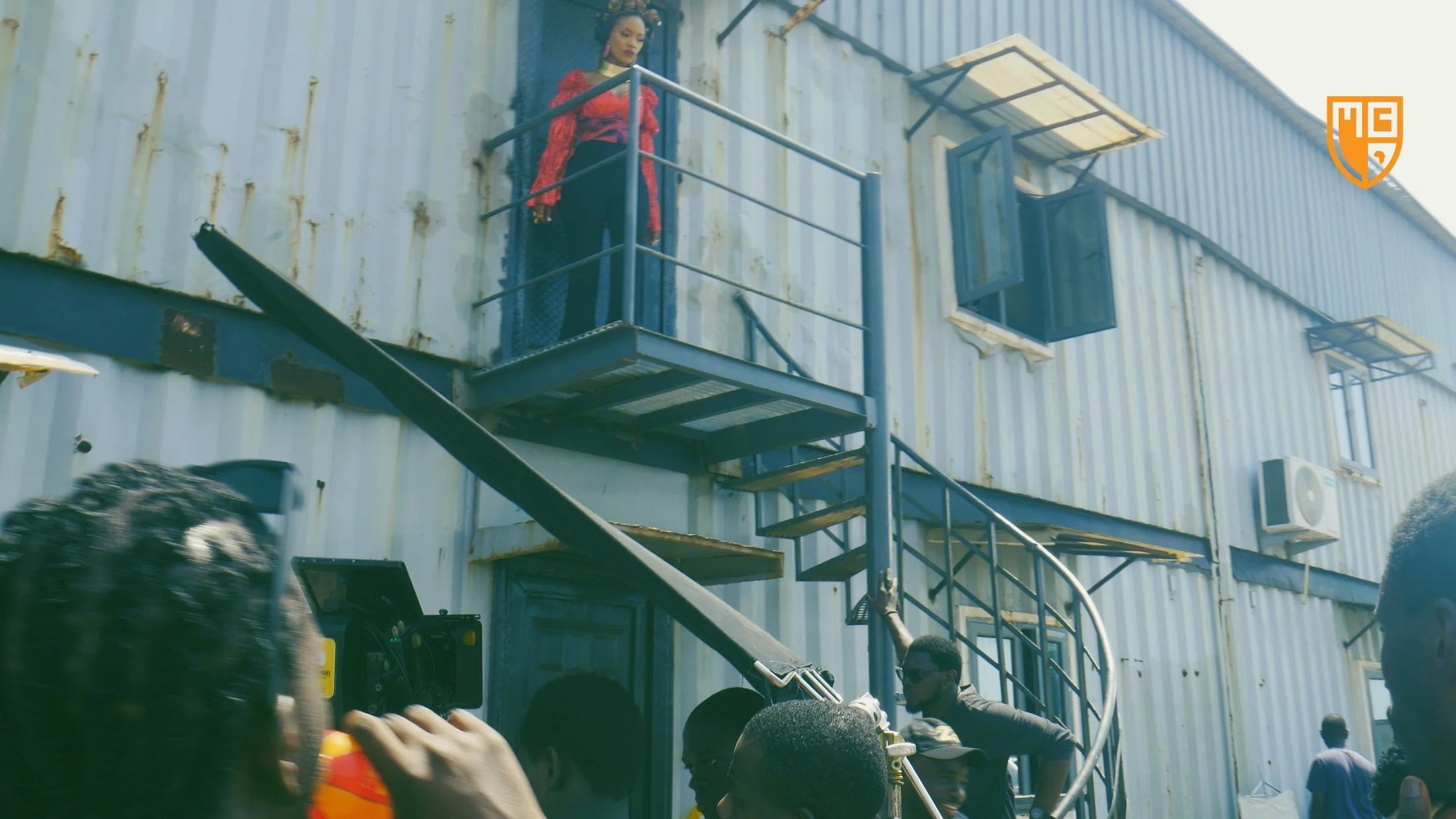 Wide behind-the-scenes image of an outdoor industrial filming location with crew, camera rig, and talent positioned on set